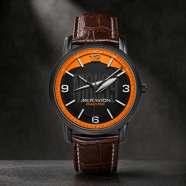 A luxury wristwatch by McRavion Grand Prix, featuring a dark gray case, a black dial with a vibrant orange inner ring and white numerals, and a brown crocodile-embossed leather strap.