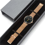 A rose gold mesh watch with a black dial and gold-tone markers, presented inside an open black gift box next to its closed lid on a white background.