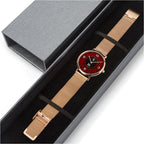 A wristwatch with a rose gold mesh strap and a red dial, presented inside a black gift box next to its gray outer packaging.