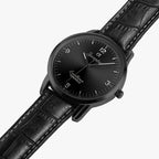 A black analog watch with a black leather strap, black round case, and a black dial featuring white Arabic numerals and hands, shown on a light gray background.