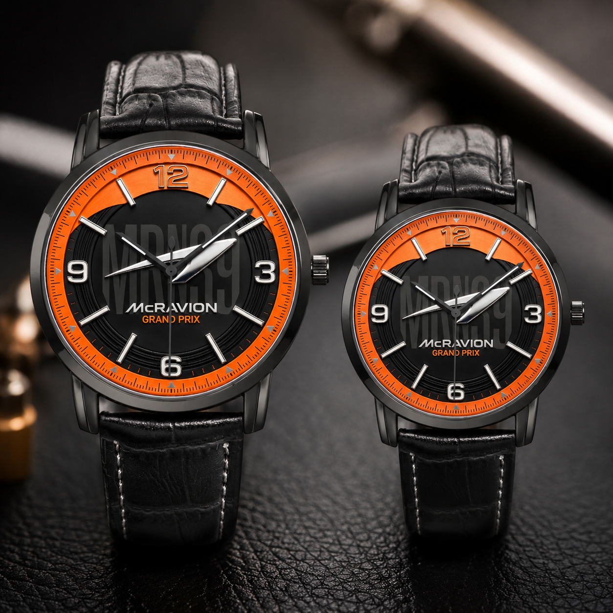 Two matching wristwatches with black leather straps and black dials featuring bright orange bezels and white numerals. The larger watch is in front of the smaller one, both branded MCRAVION GRAND PRIX, set against a dark textured surface.