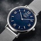 A close-up shot of a Tag Heuer watch with a deep blue dial, silver-tone stainless steel case, and matching mesh strap, set against a dark textured background.