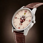 A close-up studio shot of a Ossoodo watch with a silver stainless steel case, a cream-colored dial featuring red numerals and a central Ferrari-style shield logo, and a brown leather strap against a dark brown gradient background.