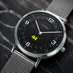 A Petronas wristwatch with a silver mesh strap, round silver case, and a black dial featuring white Arabic numerals and a bright yellow '44' in the center, shown on a dark textured background.