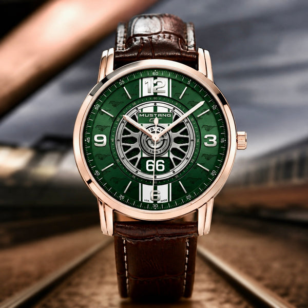 A luxury men's analog watch with a green dial, rose gold case, and brown leather strap, positioned on train tracks with a blurred train and landscape in the background.