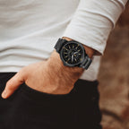 A close-up of a person's wrist wearing a black smartwatch with a black strap and gold-tone details, paired with a white long-sleeve shirt and black pants.
