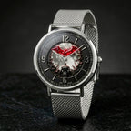 A silver-tone metal mesh strap watch with a round black dial featuring a red bat logo and a dark illustrated center, photographed on a dark studio background.