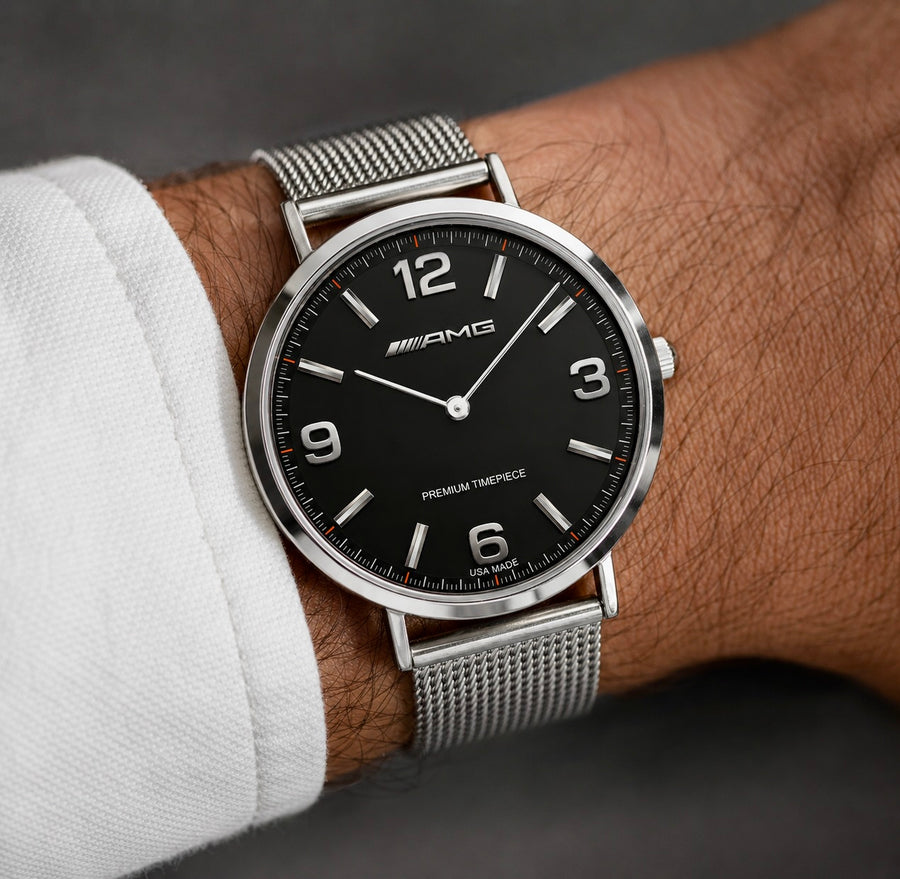 A close-up of a Mercedes-AMG watch on a person's wrist, featuring a black dial with silver-tone hands and luminous markers, a silver-tone mesh strap, and a white shirt cuff visible at the side.