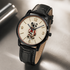 A wristwatch with a black leather strap, black case, and a cream-colored dial featuring a cartoon panda character. The watch is photographed on a reflective metallic surface.