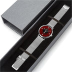 A silver mesh strap watch with a round red dial and black hands, presented diagonally inside a dark gray gift box with its lid placed above.
