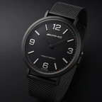 A black AMG analog wristwatch with a black mesh strap, black dial, and silver-tone numerals, shown on a dark background.