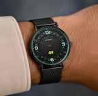 A close-up of a wrist wearing a Petronas smartwatch with a black mesh strap and a black watch face featuring white numerals and a yellow digital time display.