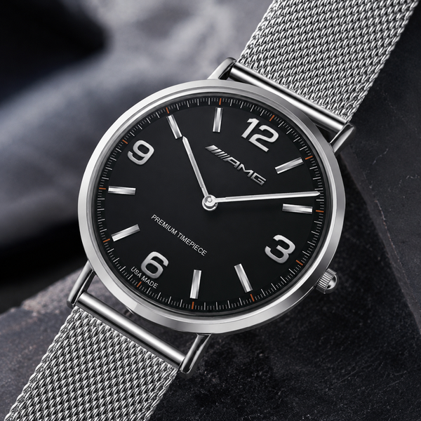 A close-up of a silver-tone metal mesh watch with a black dial, silver bezel, and silver mesh strap, resting on a dark textured surface.