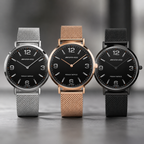 Three Mercedes-Benz wristwatches with black dials and mesh straps are lined up on a gray surface. The watches have silver, rose gold, and black metal cases and mesh bands.